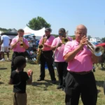 Local brass musicians performing among attendees and families at the Willingboro Jazz Festival on a sunny summer afternoon.