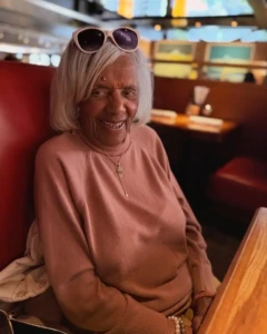 Sit With It: Willingboro | No Soldier Left Behind 2 Elderly woman smiling in a diner booth, wearing a sweater and sunglasses on her head.